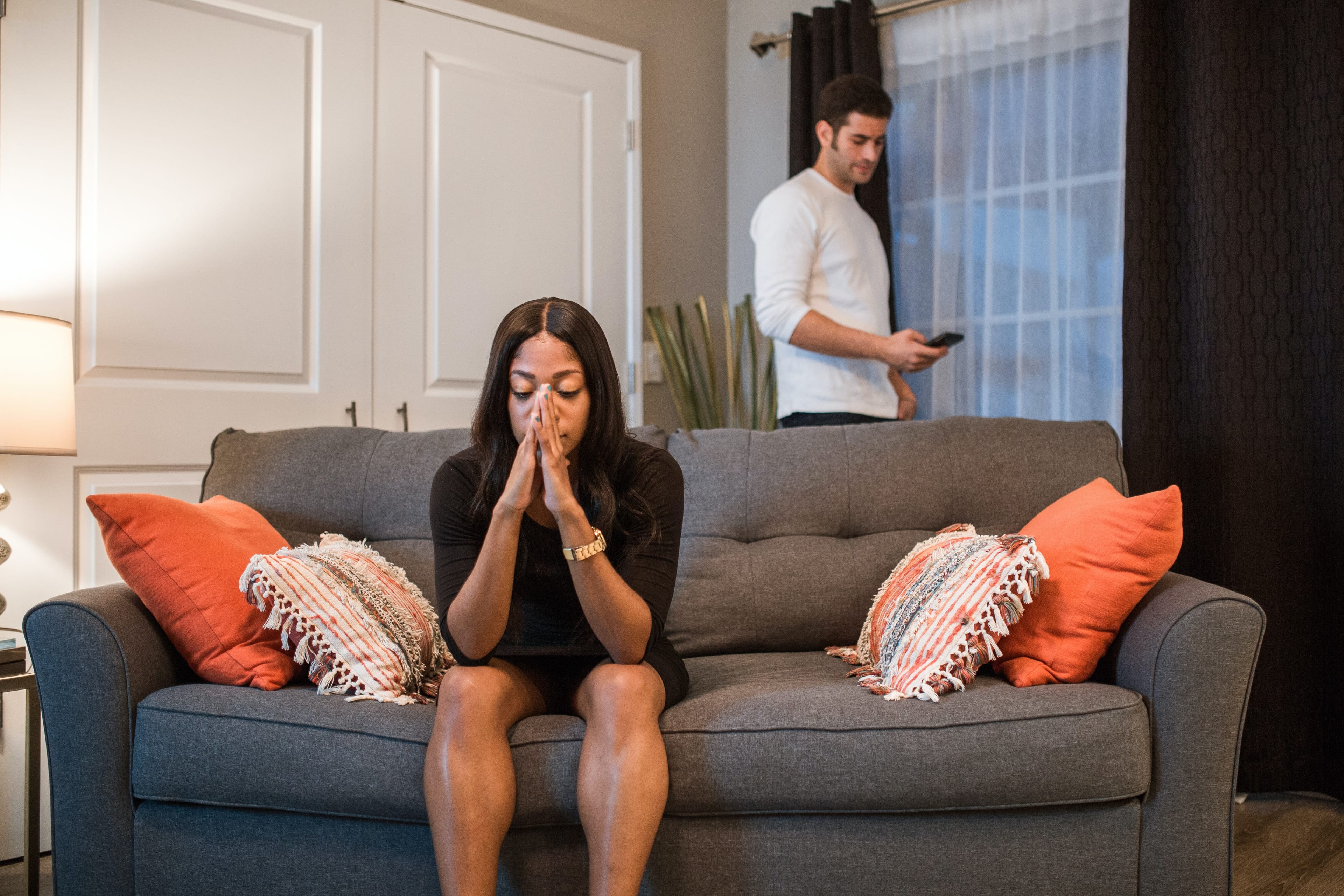 Woman sitting on a couch with her head in her hands, while a man (her partner) stands behind the couch looking at his phone