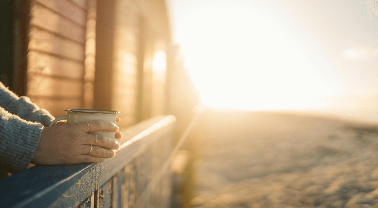 Person leaning on a balcony railing, holding a coffee mug, with a sunrise in the background