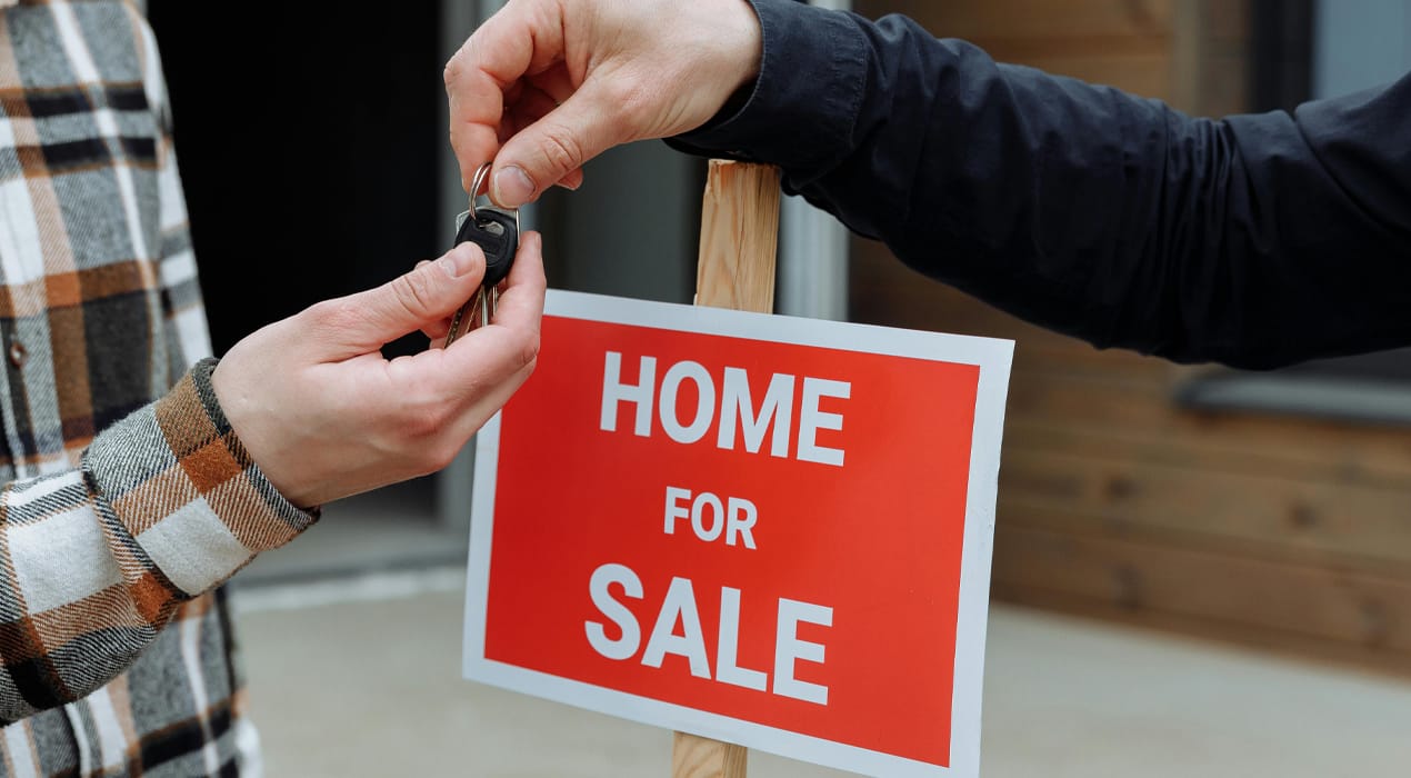 Two people standing in front of a 'home for sale' sign, with one passing the other a set of keys
