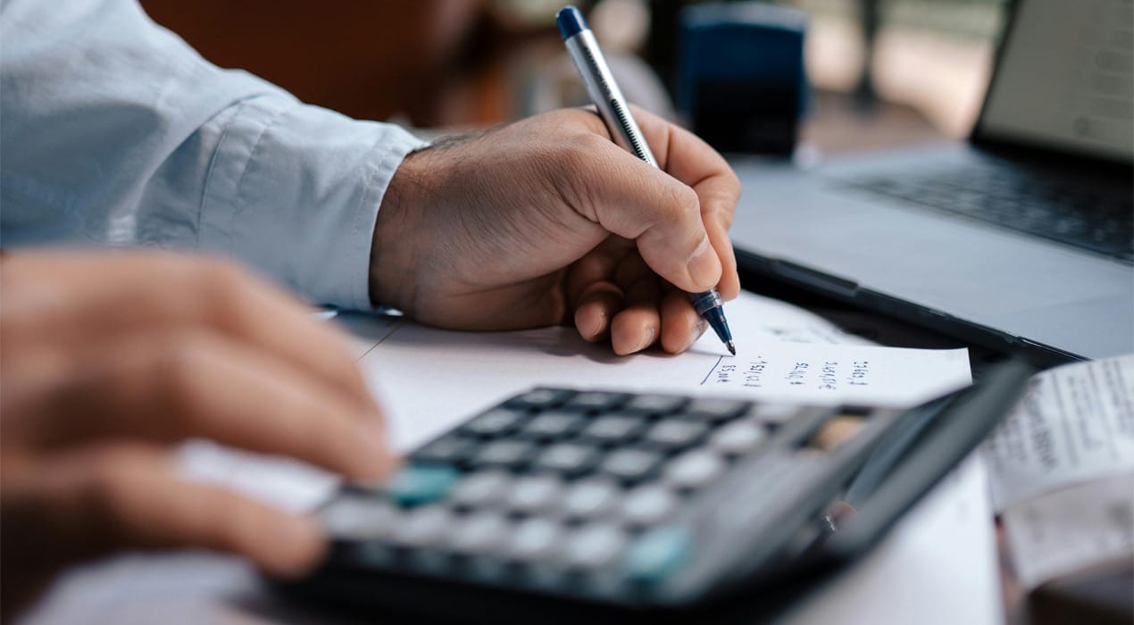 Close up of someones hands at a desk—writing with one and using a calculator with the other