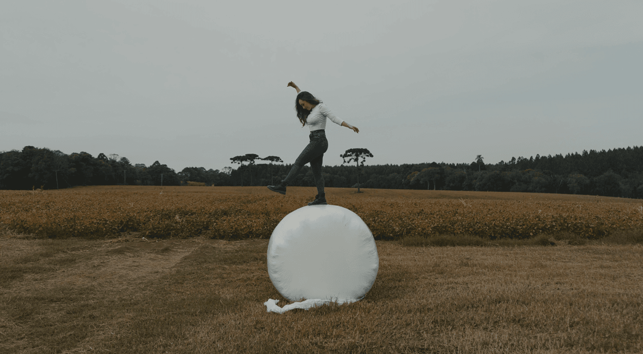 Woman balancing on top of a wrapped bale of hay, with a field and forest in the background