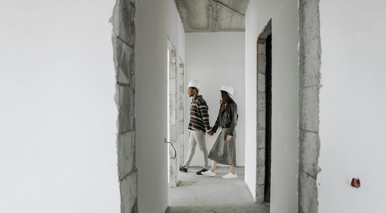 Couple walking hand in hand, wearing hard hats, through a home under construction.